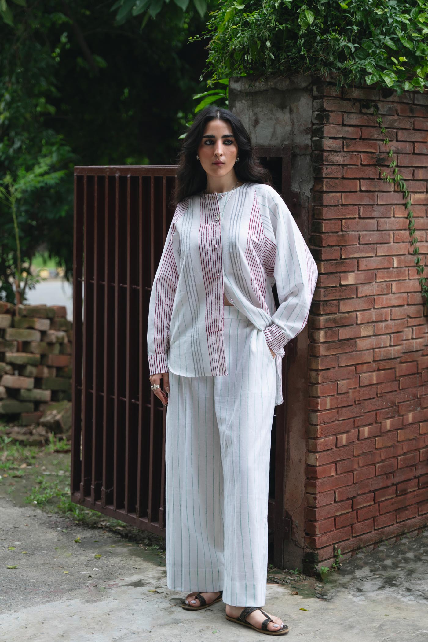 Woman in a white outfit standing against a brick wall with greenery in the background