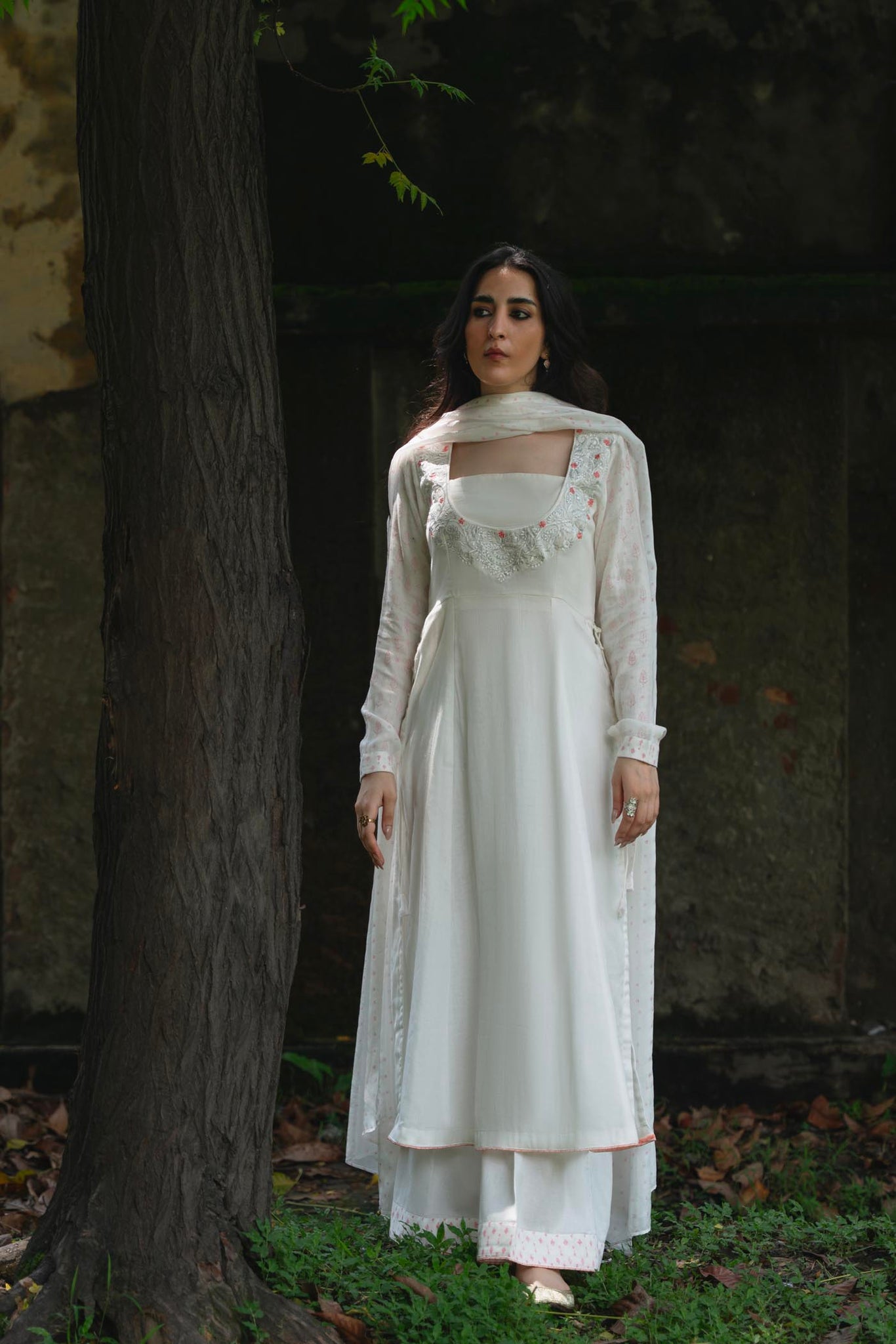 Woman in a white dress standing against a dark wall with a tree trunk.