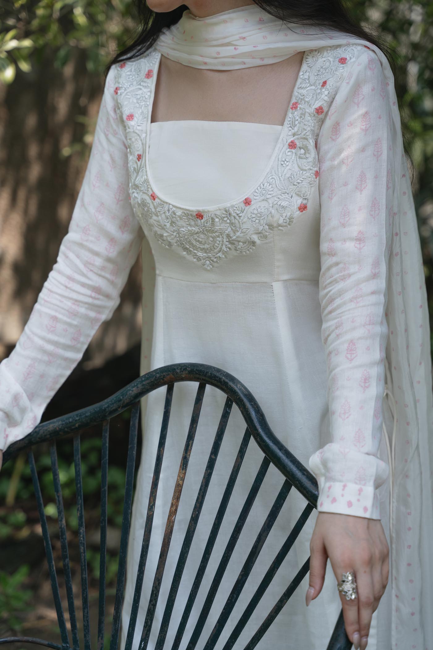 Woman wearing a white embroidered dress with a sheer overlay sitting on a chair outdoors.