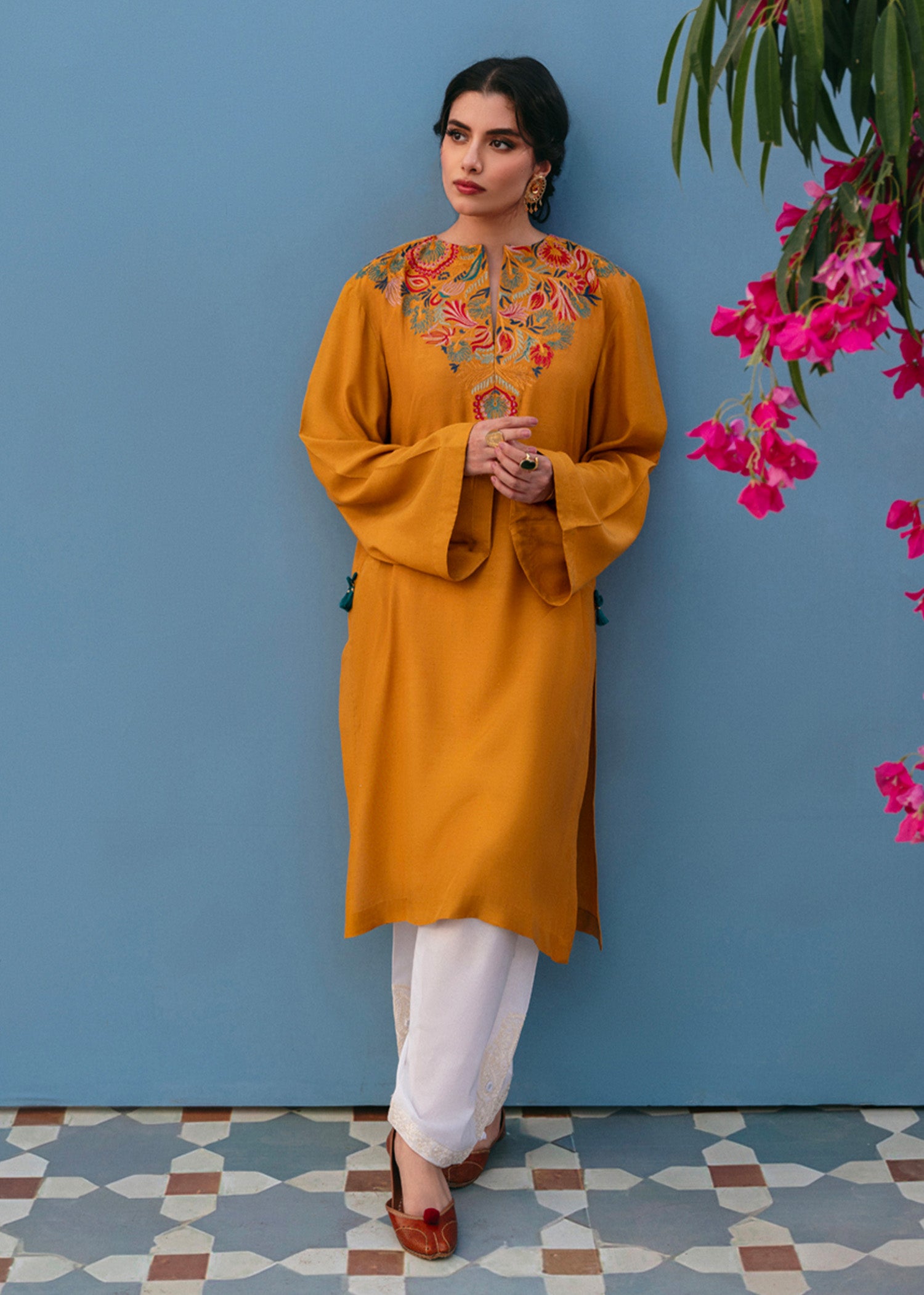 A woman wearing a mustard yellow kurta set with embroidery and tassel details, paired with white trousers, standing against a blue background with pink flowers in the background.